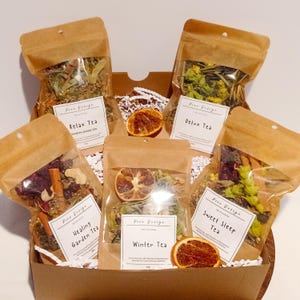 Può includere: Una scatola regalo piena di sei piccoli sacchetti di carta marrone contenenti miscele di tè sfuso. I sacchetti sono etichettati con i nomi dei tè: Relax Tea, Detox Tea, Healing Garden Tea, Winter Tea, Sweet Sleep Tea.