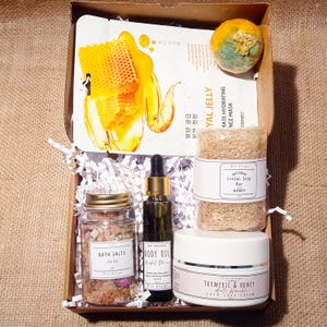 Puede incluir: Una caja de regalo llena de productos para el baño y el cuerpo, que incluyen sales de baño, aceite corporal, una barra de jabón de miel, una mascarilla facial y una crema facial. La caja está forrada con papel de seda blanco y tiene un fondo de arpillera.