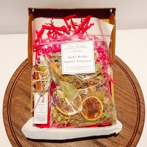 Könnte beinhalten: Eine durchsichtige Plastiktüte gefüllt mit einem süßen Winter-Simmer-Potpourri. Die Tüte ist mit "Sweet Winter Simmer Potpourri" beschriftet und befindet sich in einer rosa-weißen Geschenkbox.