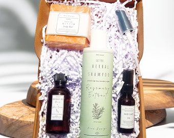 May include: A gift box filled with natural hair care products. The box contains a bar of soap, a bottle of rosemary extract shampoo, a bottle of hair oil, and a spray bottle. The products are all in a natural brown box with white shredded paper.