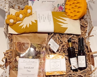 May include: A gift box filled with self-care items, including a yellow eye pillow, a yellow scalp brush, a bottle of body oil, a bottle of hair care oil, a bar of soap, a bag of tea, and a small glass jar of bath salts. The box is lined with brown crinkled paper and has a gold polka dot pattern.