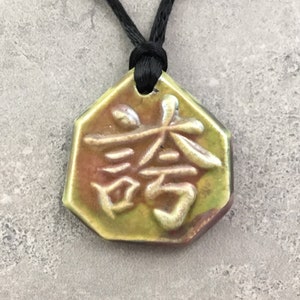 May include: A ceramic pendant with a black cord. The pendant is an octagon shape with a green and brown glaze. It features white embossed Asian characters. The cord is knotted at the top.