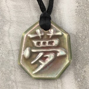 May include: A ceramic pendant with a Japanese character design, suspended from a black cord. The pendant is an irregular octagon shape with a glossy, iridescent finish in shades of green, brown, and pink. The Japanese character is raised and in a contrasting color.