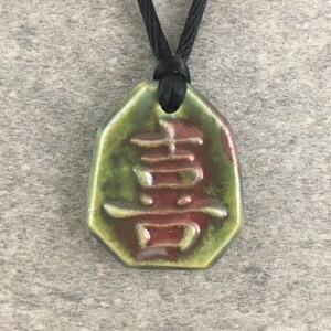 May include: A ceramic pendant with a green and maroon glaze, featuring a Chinese character. The pendant is octagonal and hangs from a black cord. The character is embossed and in a maroon color.
