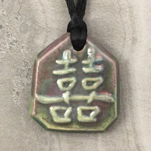 May include: A ceramic pendant with a double happiness symbol in light green against a background of green, red, and brown. The pendant is hexagonal and hangs from a black cord.