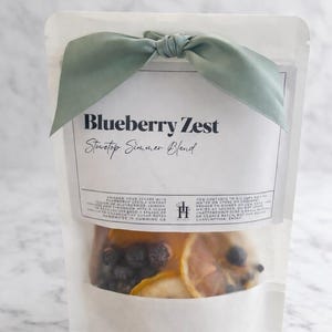 May include: A clear pouch containing a stovetop simmer blend, tied with a sage green ribbon. The label reads "Blueberry Zest Stovetop Simmer Blend." The pouch contains blueberries and lemon slices.