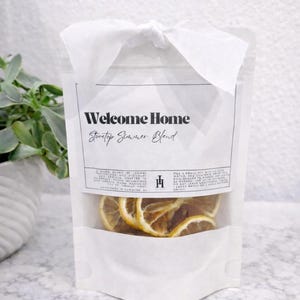May include: A white pouch with a clear window, tied with a white ribbon. The pouch contains dried lemon slices. Text on the pouch reads "Welcome Home Stovetop Summer Blend".