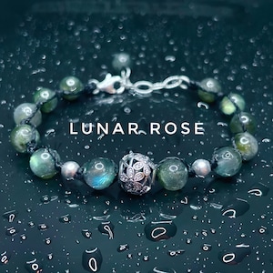 Pu&ograve; includere: Un braccialetto di perline verdi e argentate. Il braccialetto presenta una chiusura argentata e una perlina decorativa argentata con un motivo floreale. Il testo "LUNAR ROSE" &egrave; visualizzato sull'immagine.