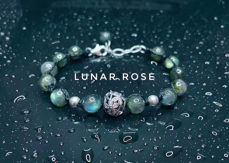 Pu&ograve; includere: Un braccialetto di perline verdi e argentate. Il braccialetto presenta una chiusura argentata e una perlina decorativa argentata con un motivo floreale. Il testo "LUNAR ROSE" &egrave; visualizzato sull'immagine.