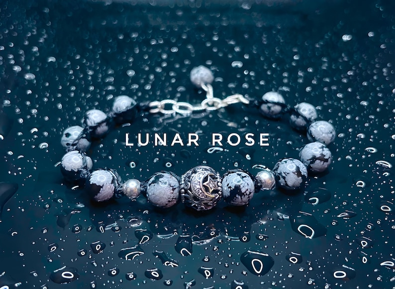 May include: A silver bracelet with a moon and star charm, featuring black and white speckled beads. The bracelet is on a dark blue background with water droplets.