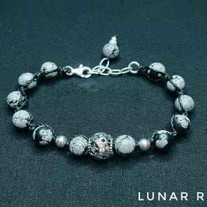 May include: A black and white stone bracelet with a silver clasp and a silver bead in the center. The bracelet is made of round beads with a black and white pattern.