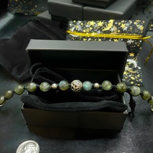 Pu&ograve; includere: Un braccialetto di perline con pietre verdi e grigio-blu e una perlina in filigrana argentata &egrave; esposto in una scatola regalo nera aperta. Il braccialetto &egrave; su un cordoncino nero. Altre scatole regalo nere e una moneta sono sullo sfondo.