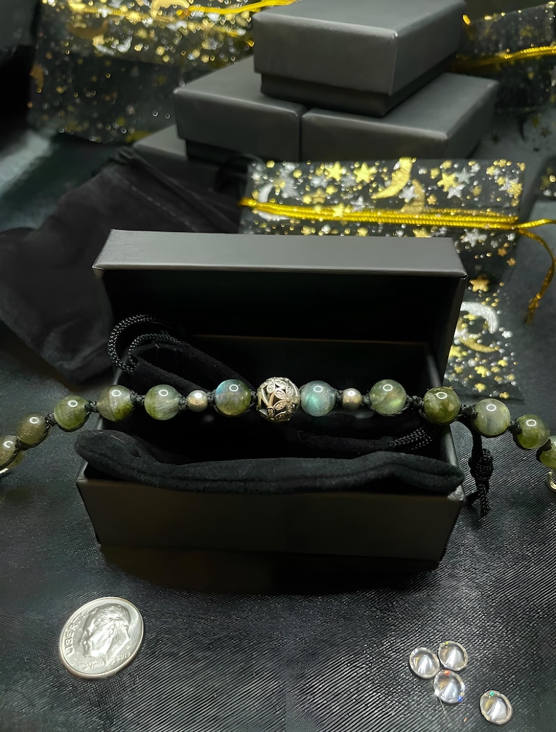 Pu&ograve; includere: Un braccialetto di perline con pietre verdi e grigio-blu e una perlina in filigrana argentata &egrave; esposto in una scatola regalo nera aperta. Il braccialetto &egrave; su un cordoncino nero. Altre scatole regalo nere e una moneta sono sullo sfondo.