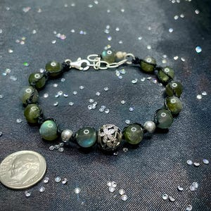 Pu&ograve; includere: Un braccialetto di perline con pietre di labradorite verdi, perline d'argento e una perlina decorativa d'argento. Il braccialetto &egrave; infilato su un cordoncino nero e ha una chiusura d'argento.