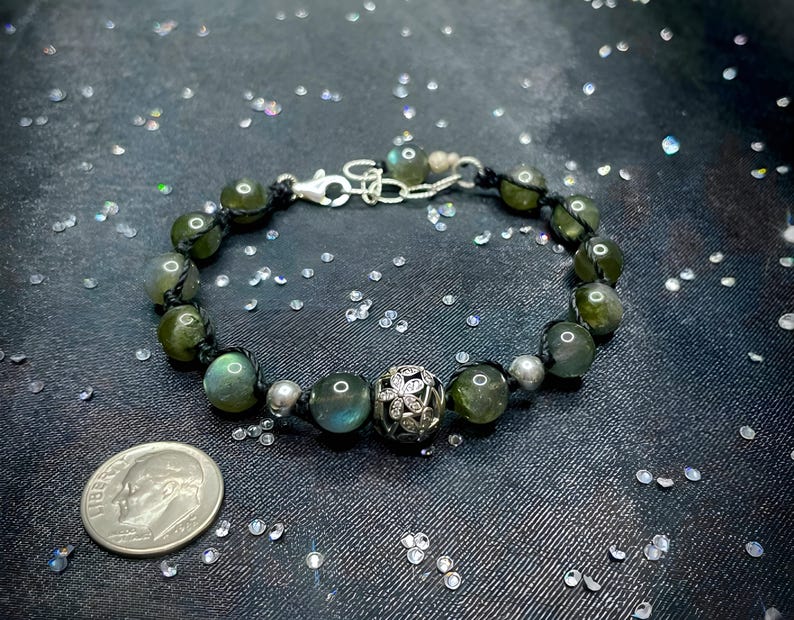 Pu&ograve; includere: Un braccialetto di perline con pietre di labradorite verdi, perline d'argento e una perlina decorativa d'argento. Il braccialetto &egrave; infilato su un cordoncino nero e ha una chiusura d'argento.