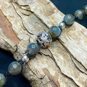 Pu&ograve; includere: Un braccialetto di perline con pietre di labradorite grigio-verdi iridescenti e dettagli argentati. Il braccialetto include una perlina decorativa argentata con un motivo floreale. Le perline sono infilate su un cordoncino nero.