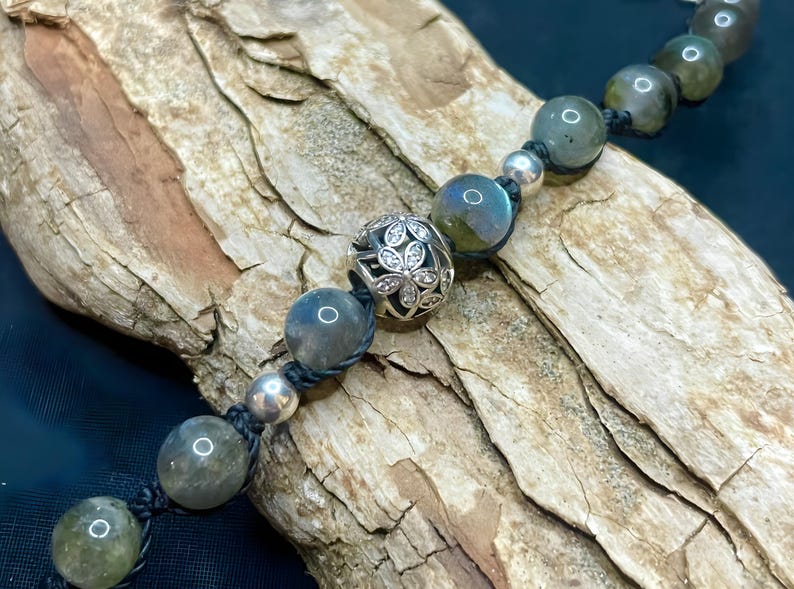 Pu&ograve; includere: Un braccialetto di perline con pietre di labradorite grigio-verdi iridescenti e dettagli argentati. Il braccialetto include una perlina decorativa argentata con un motivo floreale. Le perline sono infilate su un cordoncino nero.