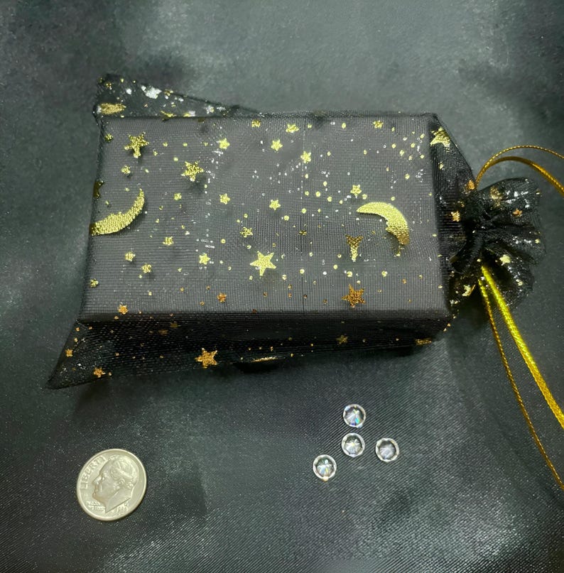 May include: A small black box with a gold star and crescent moon pattern wrapped in a black sheer fabric with gold stars and moons. The box is sitting on a black surface with a few clear rhinestones scattered around it.