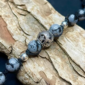 May include: A black cord bracelet with grey and white snowflake obsidian beads and a silver charm with a star and crescent moon design.