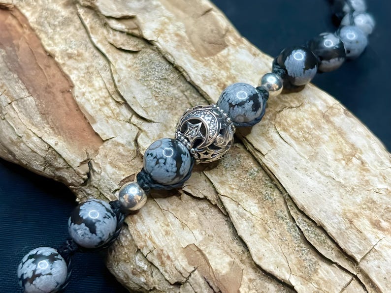 May include: A black cord bracelet with grey and white snowflake obsidian beads and a silver charm with a star and crescent moon design.