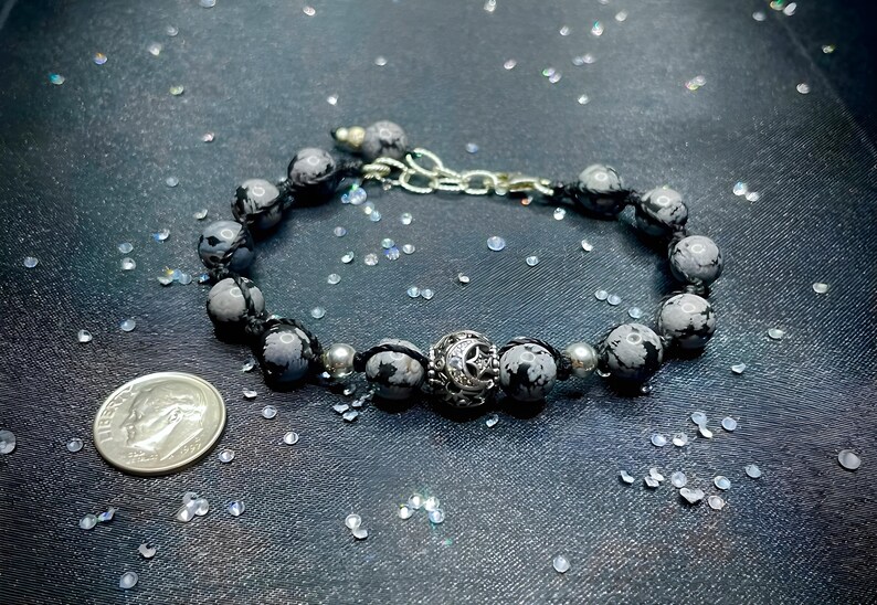 May include: A black and white stone bracelet with a silver charm featuring a crescent moon and star. The bracelet is made with black cord and silver beads.