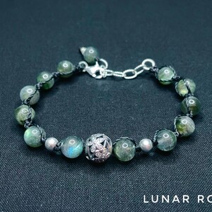Pu&ograve; includere: Bracciale con perline verdi e argentate, con un ciondolo decorativo argentato. Il bracciale &egrave; infilato su un cordino nero e ha una chiusura argentata. Il testo "LUNAR ROSE" &egrave; in basso.