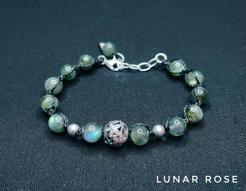 Pu&ograve; includere: Bracciale con perline verdi e argentate, con un ciondolo decorativo argentato. Il bracciale &egrave; infilato su un cordino nero e ha una chiusura argentata. Il testo "LUNAR ROSE" &egrave; in basso.