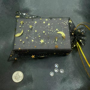 Pu&ograve; includere: Una scatola regalo nera con stelle e lune dorate &egrave; avvolta in un sacchetto di rete nero con un cordoncino dorato. Un dime americano e quattro piccole gemme rotonde e trasparenti sono visibili.