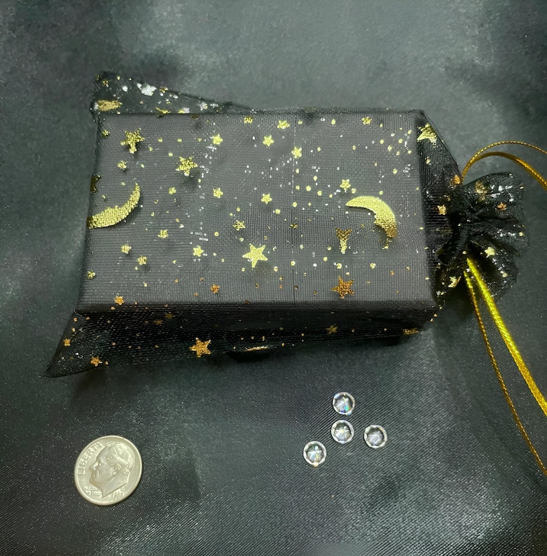 Pu&ograve; includere: Una scatola regalo nera con stelle e lune dorate &egrave; avvolta in un sacchetto di rete nero con un cordoncino dorato. Un dime americano e quattro piccole gemme rotonde e trasparenti sono visibili.