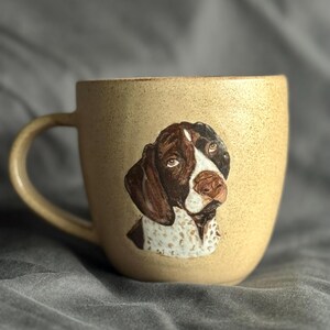 May include: A beige ceramic mug with a detailed dog portrait. The dog has brown and white markings, with a speckled pattern on its chest. The mug has a rounded shape and a matching beige handle.