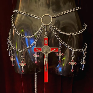 Pode incluir: Um cinto de corrente prateada com várias camadas e pingentes. Os pingentes são pequenas cruzes com esmalte vermelho e um crucifixo maior com uma figura de Jesus de prata.