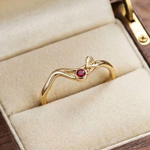 May include: A gold ring featuring a delicate wave-shaped band and a small, round, deep red gemstone set at the center. The ring is displayed inside a dark brown wooden box with a cream-colored velvet interior lining and gold-toned metal hinges.
