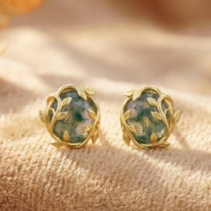 Puede incluir: Un par de pendientes de botón dorados. Cada pendiente presenta una piedra verde redonda con un patrón único, enmarcada por delicados adornos en forma de hojas. Los pendientes están colocados sobre un fondo neutro y texturizado, con hojas de otoño.