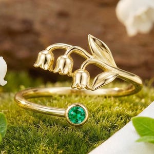 以下が含まれることがあります： スズランのデザインと小さな丸いエメラルドグリーンの宝石が付いたゴールドカラーのリング。リングは緑の苔の上に置かれ、背景には白い花と白い布があります。
