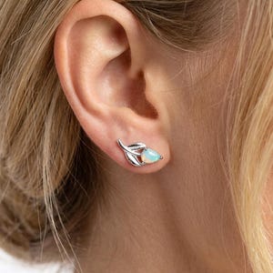 May include: A close-up of a silver stud earring in the shape of a leaf with a pear-shaped opal gemstone. The earring is worn on the ear of a person with blonde hair. The opal has a milky white color with hints of blue.