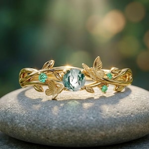 Op de afbeelding: Een goudkleurige ring met een centrale, ronde, groene edelsteen. De ring is versierd met vlinder- en bladerenmotieven en kleinere groene edelstenen. De ring wordt gepresenteerd op een grijze steen.