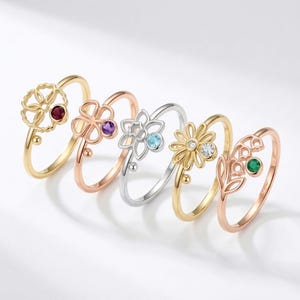 Könnte beinhalten: Fünf blütenförmige Ringe in Gold, Roségold und Silber. Jeder Ring hat einen anderen Edelstein: rot, lila, blau, klar und grün. Die Ringe werden auf einer weißen Oberfläche präsentiert.
