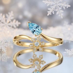 Puede incluir: Un anillo dorado con una gema azul claro en forma de pera y un diseño de copo de nieve. La banda del anillo tiene una forma fluida, y el copo de nieve está adornado con pequeñas piedras brillantes. Fondo borroso con temática invernal.