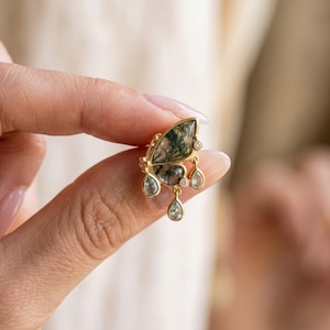 以下が含まれることがあります： 緑色のインクルージョンが入ったモスアゲートの羽を持つ金色の蝶の形をしたイヤリング。3つの小さな雫型の水色の宝石と、金属フレームにセットされた2つの小さな丸い透明な石がアクセントになっています。
