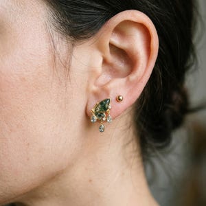 以下が含まれることがあります： 緑色のインクルージョンが入ったマーキスカットのモスアゲート石をあしらったゴールドのイヤリング。3つの小さな透明なティアドロップ型のクリスタルが揺れ、耳たぶの上部には小さな丸いスタッドピアスが付けられています。