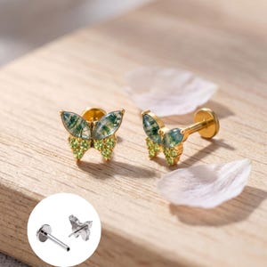 May include: A pair of gold-tone butterfly stud earrings featuring moss agate wings with green inclusions and small lime green accent stones on the lower wings. The earrings have a flat back post design, with an inset showing the silver-tone back.