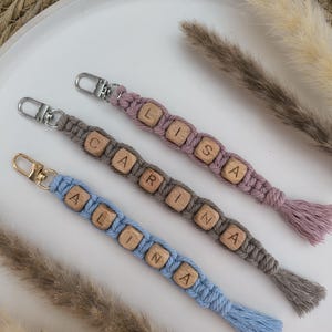 Könnte beinhalten: Drei personalisierte Makramee-Schlüsselanhänger in Pastellfarben. Jeder Schlüsselanhänger hat Holzbuchstaben, die einen Namen bilden, mit einer Quaste am Ende. Die Schlüsselanhänger sind in Blau, Grau und Lavendel gehalten, mit silber- und goldfarbenen Metallbeschlägen.