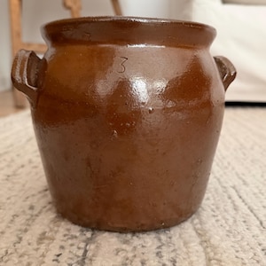May include: A brown glazed ceramic crock featuring two side handles and the number 3 embossed on the front. The vessel has a wide, rounded rim and a tapered body with a smooth, shiny finish, showing subtle surface texture and variations in color.
