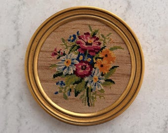 May include: A round, framed needlepoint featuring a colorful floral bouquet. The flowers include pink, blue, yellow, and white blooms with green foliage. The frame is gold-toned, and the needlepoint is set against a tan background.
