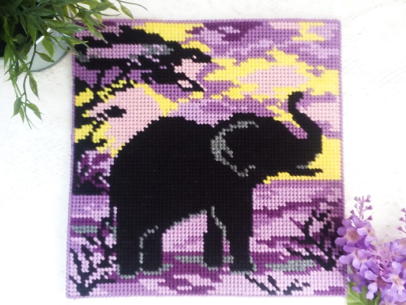 May include: A purple, yellow, and black needlepoint picture of an elephant standing in a field. The elephant is black and the field is a gradient of purple and yellow.