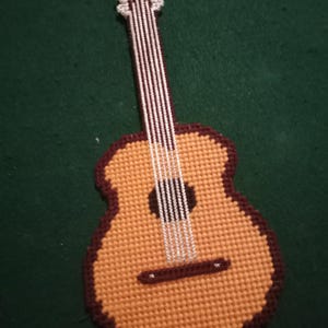 May include: A handcrafted guitar-shaped coaster. The coaster is made of tan and brown plastic canvas, with white string detailing the strings. The guitar has a dark brown outline and a sound hole. The background is a dark green.