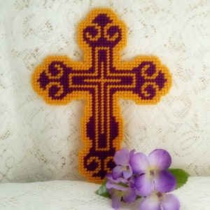 May include: A handcrafted, decorative cross in a vibrant color scheme of gold and purple. The cross features an ornate design and is made with a plastic canvas technique. A small bouquet of purple flowers rests near the cross.