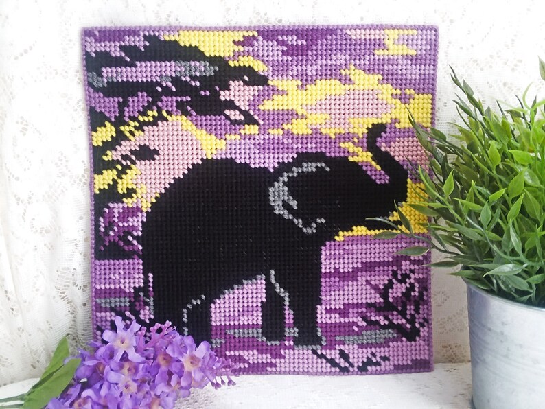 May include: A purple, yellow, and black needlepoint picture of an elephant standing in a field. The elephant is a silhouette against a sunset background.