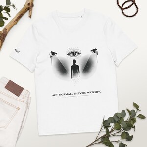 Puede incluir: Camiseta blanca con un diseño gráfico que presenta un ojo, una silueta y cámaras de vigilancia. El texto "ACT NORMAL, THEY'RE WATCHING" está impreso debajo. La camiseta es de material suave.