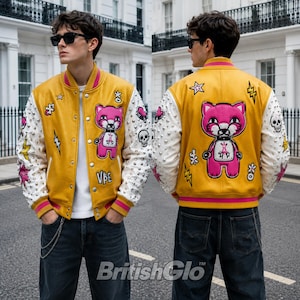 May include: A mustard yellow varsity jacket with white sleeves featuring silver studs, a pink cartoon cat graphic, and various patches including lightning bolts, skulls, and hearts. The front displays the text VBE, while the back shows a larger cat.